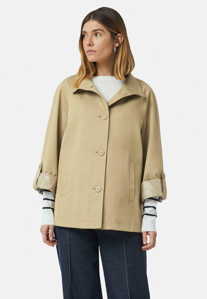 Beżowa kurtka przeciwdeszczowa z kołnierzem, guzikowym zapięciem z przodu, dwoma bocznymi kieszeniami i wzorzystymi mankietami; charakteryzuje się luźnym krojem i gładką fakturą.
