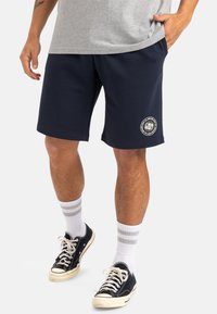 Pantalones cortos de sudadera azul marino con un ajuste relajado, confeccionados con una tela suave, que cuentan con bolsillos laterales y un diseño de logo circular en el muslo izquierdo.