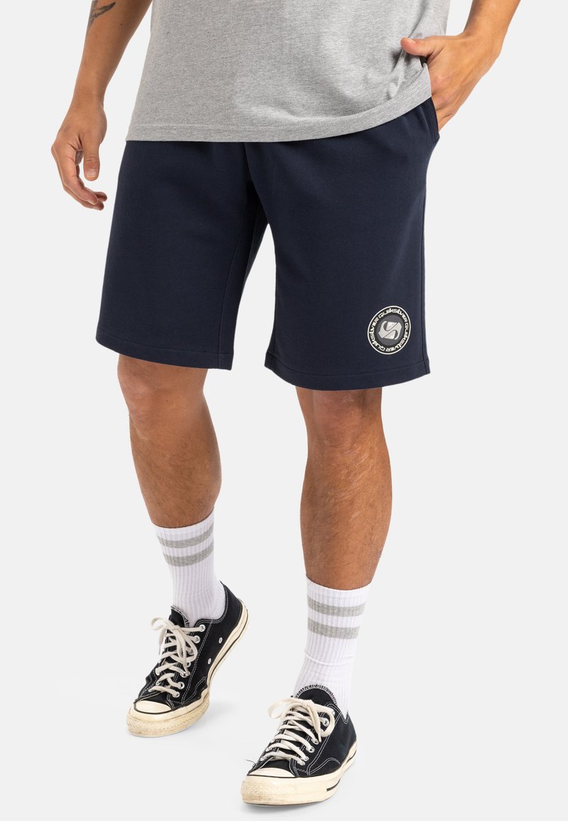 Pantalones cortos de sudadera azul marino con un ajuste relajado, confeccionados con una tela suave, que cuentan con bolsillos laterales y un diseño de logo circular en el muslo izquierdo.