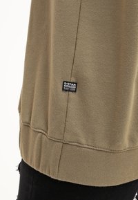 Sudadera verde oliva con puños y dobladillo acanalados, con una etiqueta negra con texto blanco. Textura suave y diseño sencillo.