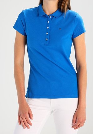 Frau in einem enganliegenden blauen Poloshirt mit kurzen Ärmeln, weißen Knöpfen und weißen Hosen, stehend vor einem hellen Hintergrund.