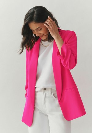 Woman with dark hair wearing bright pink blazer, white shirt, and white pants, smiling and touching her hair against a plain background.