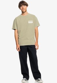 Olive green cotton t-shirt with a small white graphic on the chest, paired with dark navy pants and gray sneakers. Casual fit, short sleeves.