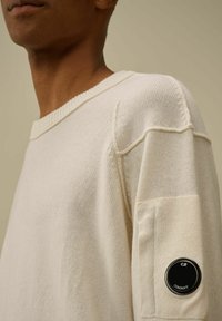 Maglione lavorato a maglia color crema con scollatura a coste e cuciture distintive. Presenta un'etichetta con logo sul maniche sinistro.