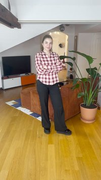 Camisa vermelha e branca aos quadrados, calças pretas, de pé num chão de madeira com um sofá castanho texturado e uma grande planta verde num vaso.