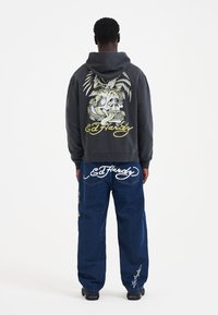 Felpa con cappuccio grigia con grande grafica colorata di teschio e aquila sul retro, abbinata a jeans blu con loghi "Ed Hardy" ricamati.