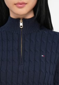 Ναυτικό πλεκτό πουλόβερ με ψηλό γιακά και χρυσό φερμουάρ, διαθέτει υφή καλωδίου και ένα μικρό λογότυπο σε κόκκινο, άσπρο και μπλε χρώμα.