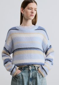 Pull rayé bleu clair en fil doux et moelleux. Présente des rayures horizontales en beige, bleu foncé et noir. Poignets côtelés.