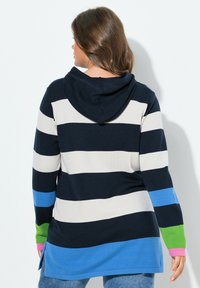 Vrouw met bruin haar draagt een hoodie met brede horizontale strepen in marineblauw, wit, blauw, groen en roze, staande tegen een witte achtergrond.
