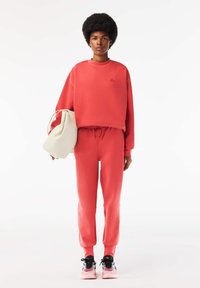 Korallfärgad sweatshirt och matchande joggingsbyxor i mjukt material, med en kortad design och elastisk midja. Modellen bär en vit väska.