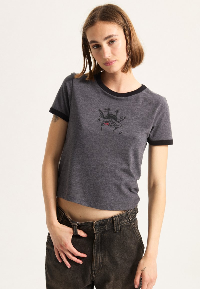 Grå beskåret t-shirt med korte sorte ærmer og et grafisk print med et skelet og stjerner på forsiden. Materiale af bomuldsblanding.