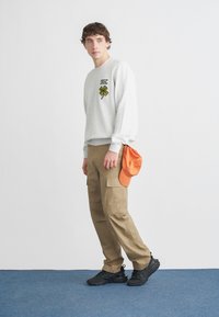 Grauer Pullover mit grünem Vierblättrigen-Kleeblatt-Design, khakifarbene Cargo-Hose, orangefarbene Mütze und schwarze Sneaker vor einem schlichten Hintergrund.