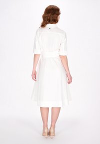 Weißes Hemdkleid mit kurzen Ärmeln, tailliertem Bund und A-Linien-Rock. Glatter Stoff mit dezentem Glanz, knielang, kombiniert mit beigen Absätzen.