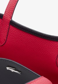 Sac à main en cuir rouge avec une surface texturée, doublure intérieure noire et détails en logo argenté. Équipé de poignées robustes et d'un design ouvert.
