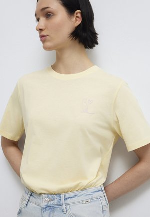 Junge Frau mit kurzen dunklen Haaren trägt ein blassgelbes T-Shirt mit einem kleinen Katzen- und Herzmotiv auf der Brust, das in eine helle Jeans gesteckt ist.