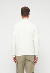 Felpa bianca con maniche lunghe e colletto alto. Presenta un piccolo logo "H" sulla parte posteriore del collo. Tessuto in cotone morbido, vestibilità rilassata.