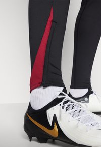Czarny zestaw sportowych legginsów z czerwoną wstawką, połączony z białymi i czarnymi korkami do piłki nożnej, wyróżniającymi się złotym logiem Nike.