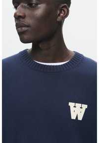 Tumedas sinises lõngast kampsun ribakääriga ümarate kaelustega ja vasakul rinnal valge tähe "W" logo. Pehme tekstuuriga ja mugavalt liibuv siluett.