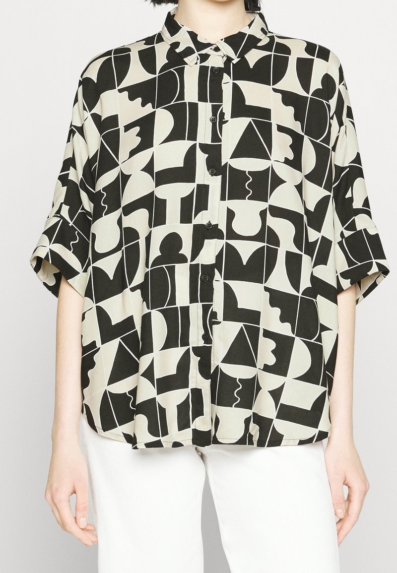 Chemise à manches courtes boutonnée avec un motif géométrique abstrait noir et blanc, portée avec un pantalon blanc.