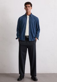 Chemise en denim à boutons de couleur bleu foncé, avec une poche poitrine. Associée à un pantalon noir ample et des chaussures noires. Fond uni.