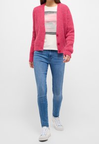 Rosa stickad cardigan med knäppning, i kombination med en grafisk t-shirt och blå skinny jeans. Modellen har på sig vita sneakers.