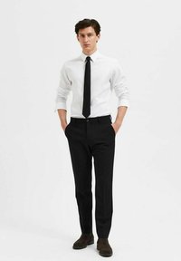 Witte overhemd met lange mouwen, zwarte stropdas en zwarte broek. De outfit heeft een slim fit met een gladde textuur en minimalistisch ontwerp, aangevuld met donkere schoenen.