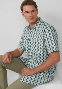 Korte mouwen, button-up shirt in lichtblauw met een palmbomenpatroon. Het materiaal lijkt lichtgewicht; gecombineerd met olijfgroene broek.