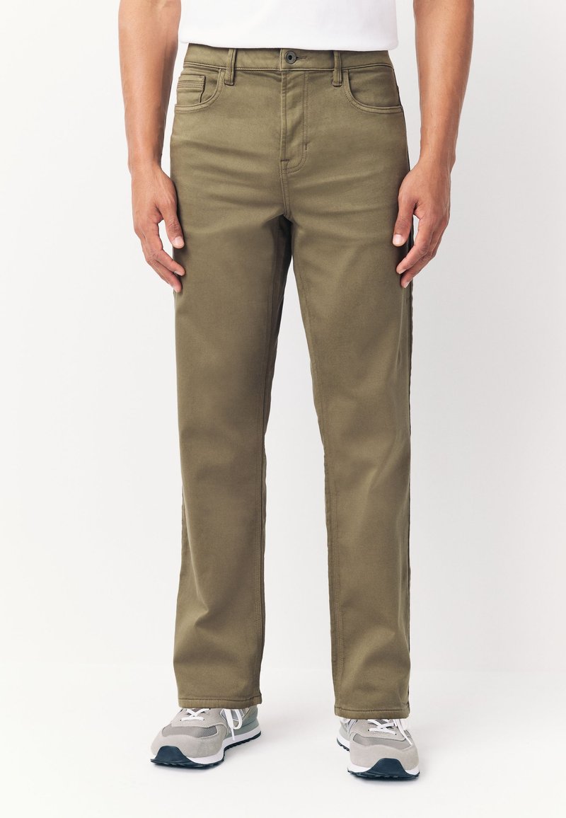 Mann trägt olivgrüne gerade Hose und graue Sneakers und steht vor einem schlichten weißen Hintergrund.
