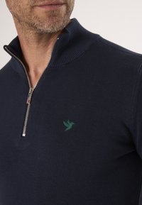 Námořnický pletený pulovr se zipovým límcem, který má na hrudi malé logo zeleného ptáčka. Hladká textura a přiléhavý design.