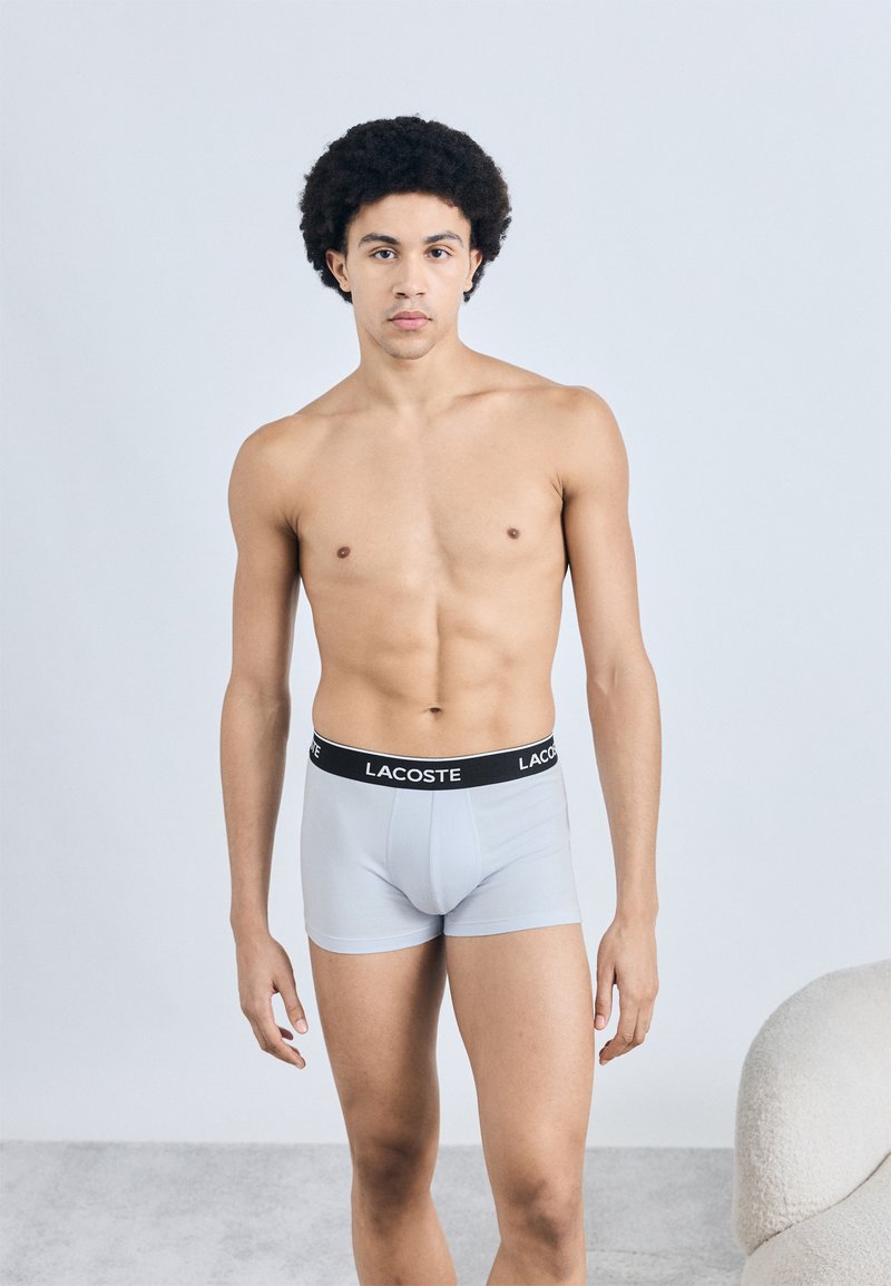 Lichtgrijze boxershorts van rekbare stof, met een zwarte tailleband met het "LACOSTE"-logo. Model staat op een neutrale achtergrond.