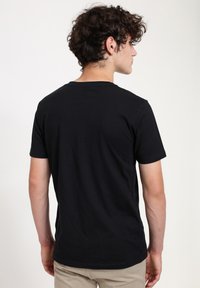 Mladý muž s kudrnatými vlasy, oblečený v jednoduchém černém tričku s krátkým rukávem a béžových kalhotách, stojící zády k jednolitému světlému pozadí.