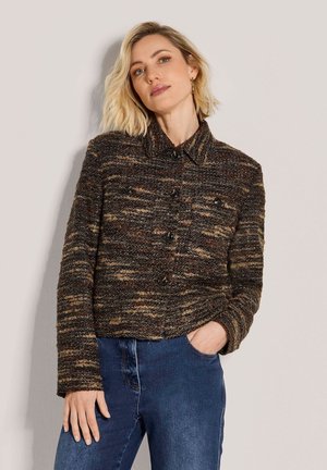 Frau mit blondem Haar trägt eine braune, strukturierte Jacke mit Knöpfen und blaue Jeans, posiert mit einer Hand in der Tasche vor einfarbigem Hintergrund.