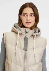 Hellbeige Puffer-Weste mit Kapuze, ausgestattet mit einem Reißverschluss, Zugband-Akzenten und glatter Textur. Graue Innenverkleidung sichtbar.