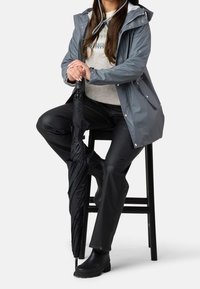 Femme assise sur un tabouret, portant un imperméable gris, un pantalon noir et des bottes, tenant un parapluie noir fermé des deux mains.