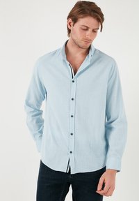 Camisa de manga larga azul clara hecha de un tejido texturizado, con un cuello puntiagudo y un cierre frontal de botones con botones oscuros. Corte casual, ligeramente metida por dentro.