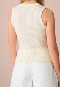 Haut en maille beige sans manches avec surface texturée, présentant une coupe ajustée et un col arrondi, associé à un pantalon blanc taille haute.