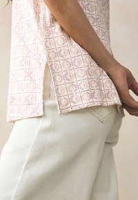 Világos rózsaszín mintás felső geometriai mintával, rövid ujjú és oldalán hasítékokkal, törtfehér nadrággal, mely hátsó zsebekkel rendelkezik.