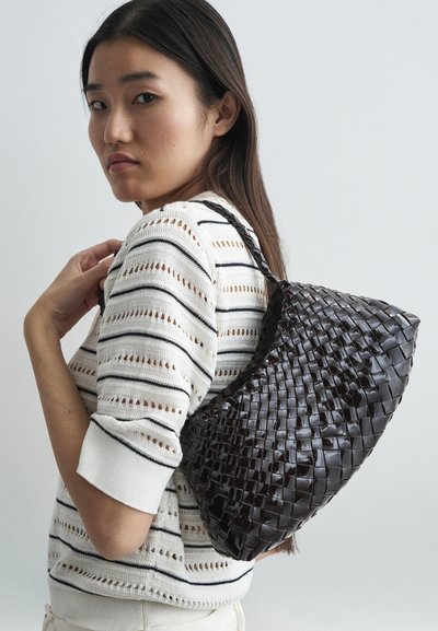 Bag de couro castanho trançado com uma forma curva e um acabamento texturizado, segurado no ombro contra um fundo claro.