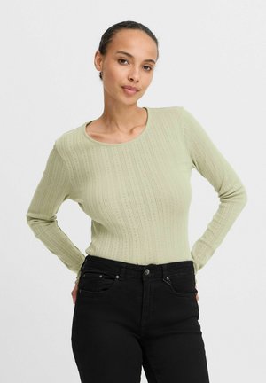 Hellgrünes langärmeliges Rippenoberteil mit Rundhalsausschnitt, kombiniert mit schwarzen, eng anliegenden Jeans, die ein strukturiertes Muster aufweisen.