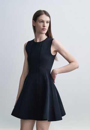 Γυναίκα με μακριά καστανά μαλλιά που φοράει αμάνικο στενό μαύρο φόρεμα με φουσκωτή φούστα, ποζάροντας με το ένα χέρι στη μέση.