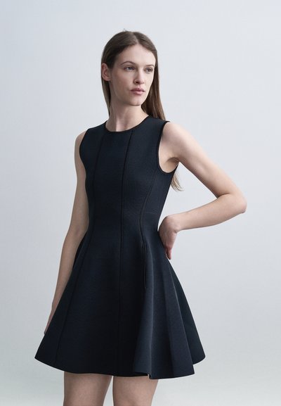 Femme aux longs cheveux bruns portant une robe noire sans manches ajustée avec une jupe évasée, posant avec une main sur la hanche.