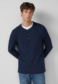 Jonge man met kroezig bruin haar, gekleed in een marineblauwe langjarige henley-shirt over een wit onderhemd en blauwe spijkerbroek, staand tegen een grijze muur.