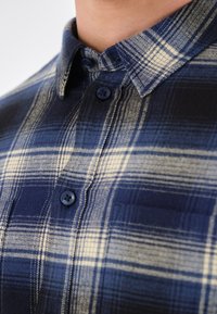 Camisa de cuadros abotonada en azul marino, crema y azul oscuro; presenta un cuello, botones oscuros y una tela suave de textura con un patrón de cuadros.