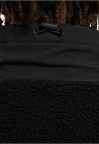 Schwarzes Fleece-Kleidungsstück mit geripptem oberen Teil und verstellbarem Kordelzug. Der untere Bereich besteht aus einem strukturierten, weichen Stoff für zusätzliche Wärme.