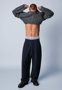 Szary puchaty sweter ściągany na tors, w połączeniu z ciemnymi spodniami. Pas ma wzór w paski, czarne buty.