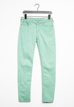 Jean slim vert menthe avec fermeture à bouton et zipper, poches avant et arrière, suspendu à un cintre noir contre un fond blanc.