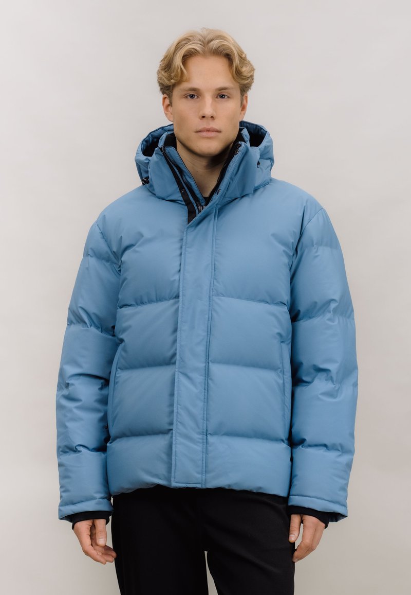 Lyseblå pufferjakke med høy krage, glidelås foran og quiltet design. Har hette og elastiske mansjetter. Stoffet ser glatt og skinnende ut.