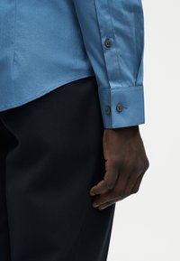 Χέρι με σκουρόχρωμο δέρμα που στηρίζεται στο πλάι, φοράει ανοιχτό μπλε πουκάμισο με μακριά μανίκια και κουμπωτά μανίκια και σκούρο παντελόνι σε ανοιχτό φόντο.