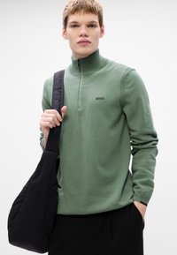 Green pullover sweater with a high collar and half-zip closure. Features ribbed accents and a printed logo on the chest. Black shoulder bag.