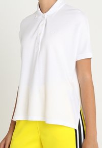 Camisa polo blanca de manga corta con cuello y tapeta de tres botones, que presenta un discreto logo en el pecho.